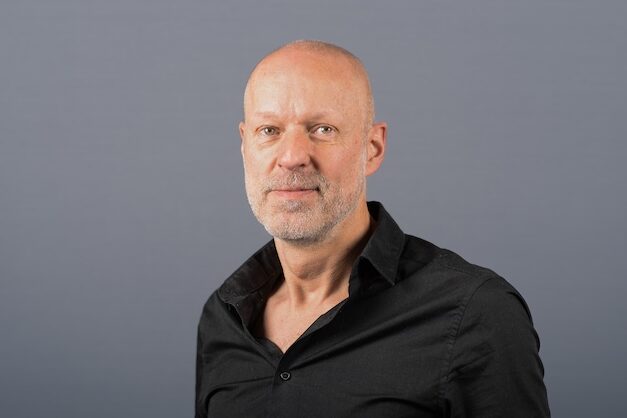 Ein Mann mittleren Alters mit rasiertem Kopf und kurzem grauem Bart, der ein schwarzes Button-up-Hemd trägt, steht vor einem schlichten grauen Hintergrund und schaut ruhig in die Kamera.