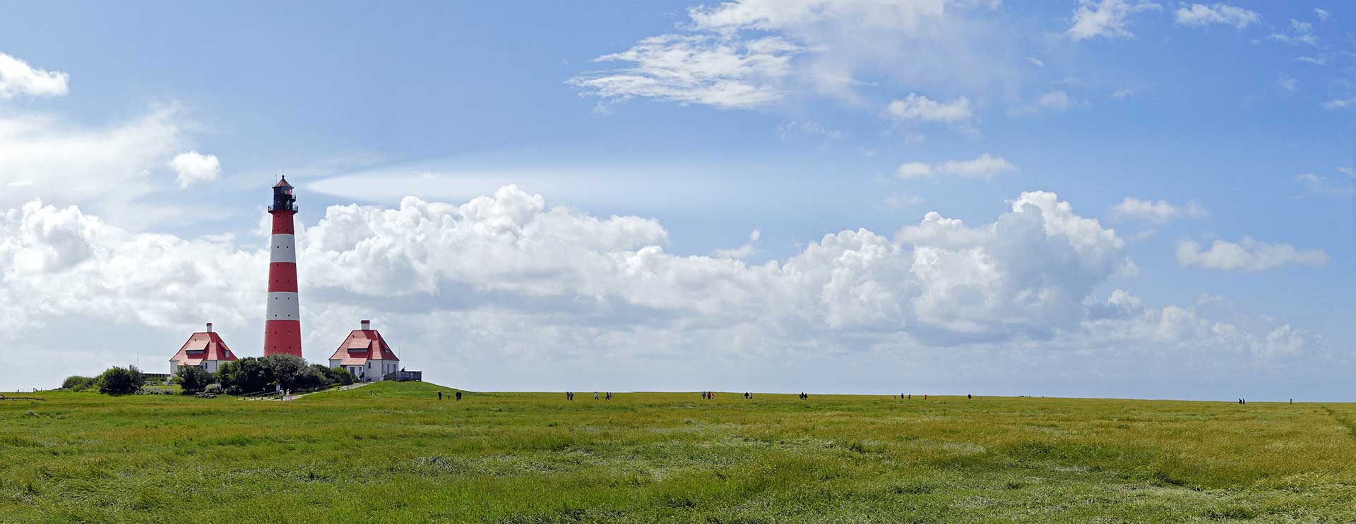 AZV Headerbild Ueber Uns Leitbild Ein rot-weiß gestreifter Leuchtturm steht auf einer grasbewachsenen Wiese unter einem blauen Himmel mit vereinzelten Wolken, flankiert von zwei kleinen Gebäuden mit roten Dächern.