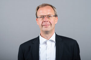 Ein Mann mittleren Alters mit Brille, einem weißen Hemd und einem schwarzen Blazer steht vor einem schlichten grauen Hintergrund und blickt mit ausdruckslosem Gesicht direkt in die Kamera.