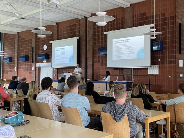 Ein Klassenzimmer oder ein Hörsaal mit Studenten, die an Tischen sitzen und einem Redner gegenübersitzen, der an einem Podium spricht. Auf zwei großen Projektionsflächen werden Folien gezeigt, und eine weitere Person sitzt an einem Tisch neben dem Podium.