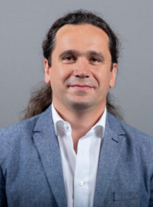 Ein Mann mit langen, nach hinten gekämmten braunen Haaren, der einen hellblauen Blazer über einem weißen Hemd trägt, steht vor einem schlichten grauen Hintergrund und blickt mit einem leichten Lächeln in die Kamera.