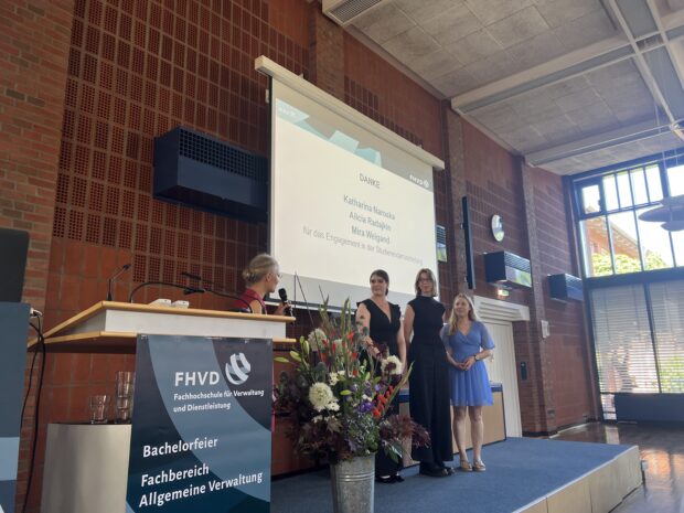 Drei Frauen stehen auf der Bühne und nehmen ihre Auszeichnung entgegen, während eine Frau vom Podium aus zu ihnen spricht. Im Bild sind eine Präsentationsfolie sowie ein Banner mit der Aufschrift „FHVD“ und „Bachelorfeier“ zu sehen. Die Szene spielt in einem sonnendurchfluteten Saal mit Backsteinwänden und großen Fenstern.