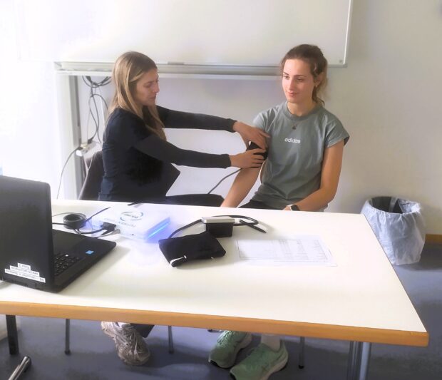 Zwei Frauen sitzen an einem Tisch; eine misst mit einer Manschette den Blutdruck der anderen. Auf dem Tisch liegen ein Laptop, medizinische Geräte und Unterlagen.